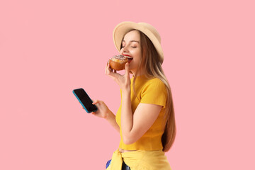 Young woman in hat with mobile phone eating tasty bun on pink background