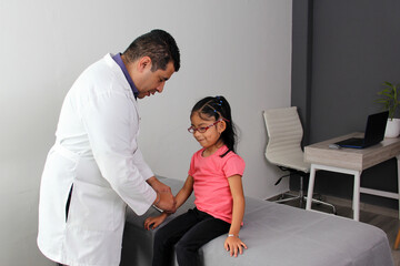 Latino pediatrician doctor attends to his 4-year-old brunette girl patient with headache, migraine and dizziness in his office wearing a white coat
