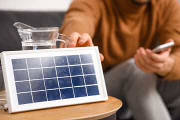Young man with mobile phone near portable solar panel at home, closeup