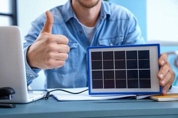 Young man charging laptop with portable solar panel in office, closeup