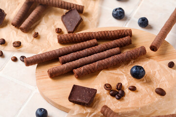 Board with delicious wafer rolls, chocolate pieces, blueberries and coffee beans on white tile background