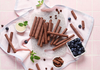 Board with delicious chocolate wafer rolls, blueberries, condensed milk and coffee beans on pink tile background