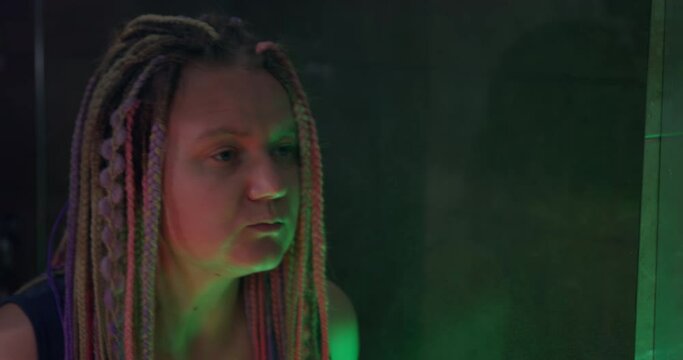 Stylish young woman with multicolored box braids hairstyle appears in the frame. She looks at herself in the mirror and explores her hair. Dim room is lit by colored lamps.