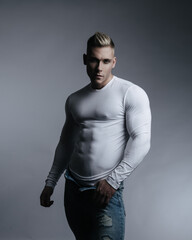 Handsome blond male model wearing white shirt and blue jeans. Sexy young man posing in tight t-shirt showing his six pack abs. Fitness guy in studio.