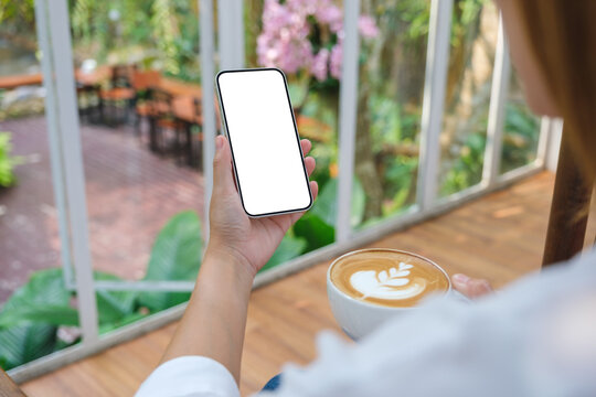 Mockup image of a woman holding and using mobile phone with blank desktop screen while drinking coffee in cafe