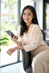Fototapeta premium Gorgeous Asian businesswoman sits on armchair, holding her smartphone, smiling and looking at camera