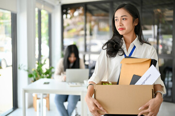 Crying Asian female office worker holding a cardboard box with her stuff, being fired from her boss - obrazy, fototapety, plakaty