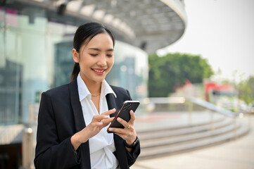 Attractive Asian businesswoman using her phone while walking outside along the company building