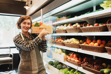 Portrait of Caucasian young adult employee is holding the basket of fruit and looking at camera with smile face. Cheerful saleswoman is working on stocking fruit on the shelves of the grocery store.
