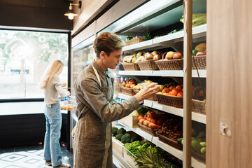 Caucasian young adult employee with an apron is looking at fruit on the shelves and checking the quality and freshness. Salesman with responsibility to quality check stock of fruit in grocery store.