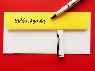 On red background, pen writing text on yellow office envelope HIDDEN AGENDA, means secret reason...