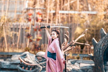 Naklejka na ściany i meble Japanese geisha in a traditional kimano with a fan and armed with a katana sword in a beautiful garden. A girl from medieval Asia. Reconstruction of cultural heritage. Culture in Japan.