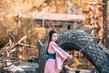 Japanese geisha in a traditional kimano with a fan and armed with a katana sword in a beautiful garden. A girl from medieval Asia. Reconstruction of cultural heritage. Culture in Japan.