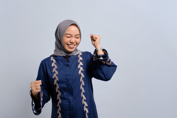 Excited young Asian Muslim woman raising fist up, celebrating success isolated over white background