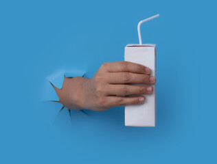 Female hand with blank packet carton juice & milk breaks through blue paper background.