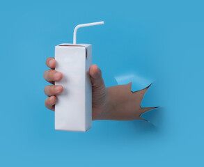 Female hand with blank packet carton juice & milk breaks through blue paper background.