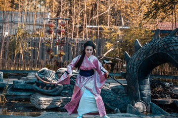 Fototapeta na wymiar Japanese geisha in a traditional kimano with a fan and armed with a katana sword in a beautiful garden. A girl from medieval Asia. Reconstruction of cultural heritage. Culture in Japan.