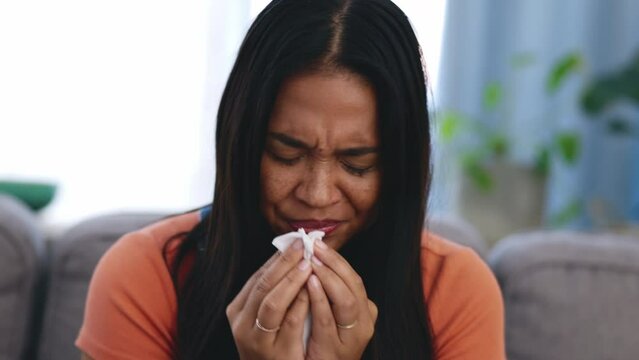 Sick, tissue and woman blowing her nose while sitting on a sofa in the living room of her home. Covid, illness and Asian female sneezing with flu, cold or allergies in the lounge of her modern house.