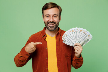 Elderly smiling man 40s years old he wears casual clothes red shirt t-shirt hold point finger on fan of cash money in dollar banknotes isolated on plain pastel light green background studio portrait.