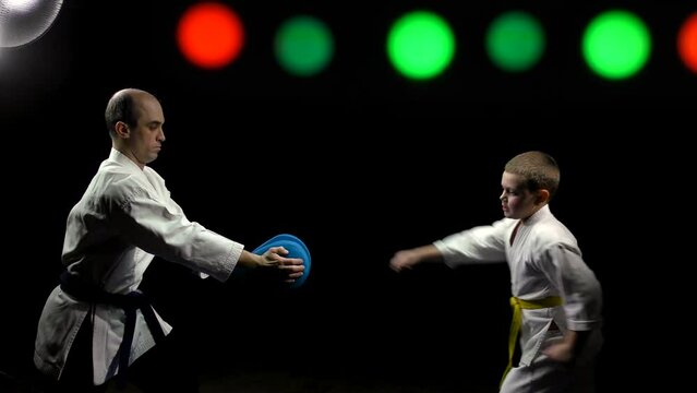 Boy athlete with a yellow belt trains kicks on the simulator on a black background with color-changing spotlights
