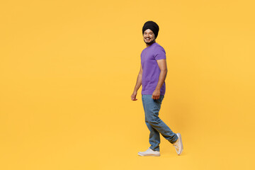 Fototapeta na wymiar Full body fun side view smiling cheerful devotee Sikh Indian man ties his traditional turban dastar wear purple t-shirt walking going look camera isolated on plain yellow background studio portrait.