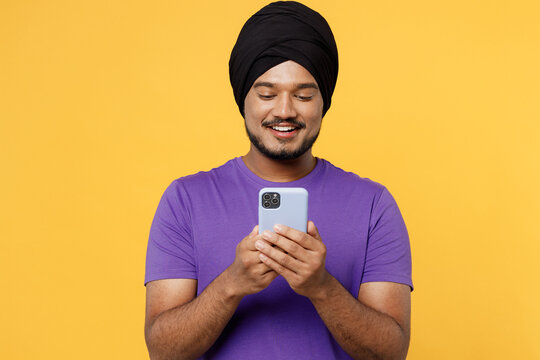 Smiling devotee Sikh Indian man ties his traditional turban dastar wear purple t-shirt hold in hand use mobile cell phone isolated on plain yellow background studio portrait. People lifestyle concept.