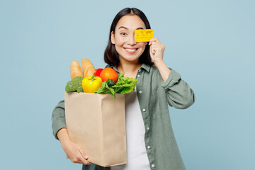Fototapeta na wymiar Young woman wear casual clothes hold brown paper bag with food products cover eye with credit bank card isolated on plain blue cyan background studio portrait Delivery service from shop or restaurant