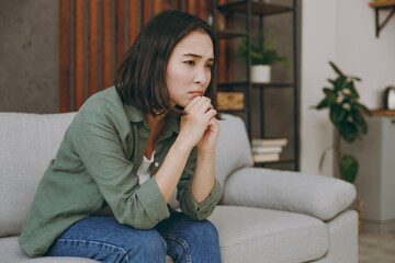Side view sad young woman of Asian ethnicity wear casual clothes prop up chin seek for solution sits on grey sofa couch stay at home hotel flat rest relax spend free spare time in living room indoor.