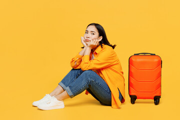 Young sad worried upset woman wear summer casual clothes sit near suitcase isolated on plain yellow...