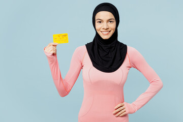 Young fun arabian asian muslim fitness trainer sporty woman wear pink abaya hijab spend time in home gym hold credit bank card isolated on plain blue background studio. Workout sport fit abs concept.