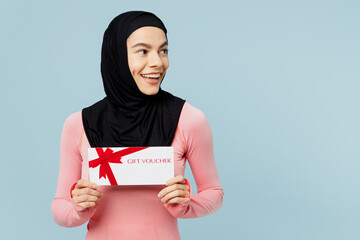 Young asian muslim fitness trainer sporty woman wear pink abaya hijab spend time in gym hold gift coupon voucher card look aside isolated on plain blue background studio Workout sport fit abs concept