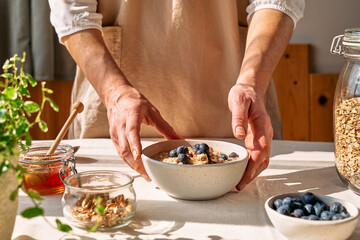 Woman preparing healthy dieting vegan nutritious breakfast. Female hands holding bowl of oatmeal porridge with blueberries, walnuts and honey.