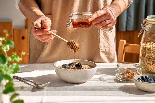Woman preparing healthy dieting vegan nutritious breakfast. Female hand pouring honey in the bowl with oatmeal porridge with walnuts and blueberries.