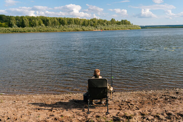 Fototapeta na wymiar Back view of unrecognizable fisherman wearing raincoat sitting on river bank on travel chair with fishing rod waiting for catch on summer sunny day. Concept of lifestyle, leisure activity on nature
