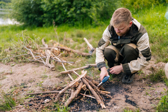 Bearded survivalist male in raincoat starting fire using gas lighter on bank of river for cooking and warming in evening before sunset. Concept of exploration, travel and adventure.
