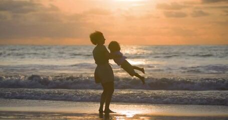 Aunt and nephew play by ocean on beach. The aunt holds and spins boy, having fun and laughing. A woman spins her nephew around while they are on the beach in the warm sunset. Games by the ocean surf.