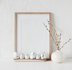 Easter background. Wooden frame mockup with copy space,spring willow branches in a vase and Easter eggs and funny bunny decor near white textured wall in neutral colors.