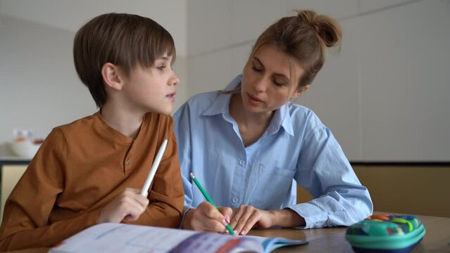 Young Caucasian mother helping child son with homework at home, sitting together at kitchen table. Happy schoolboy learning foreign language with private teacher tutor at home. Homeschooling concept