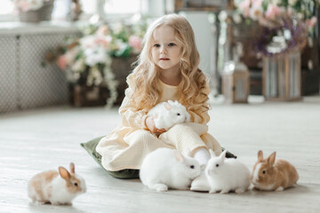 A little smiling girl in a yellow dress is playing on the floor in a room with rabbits. A baby and a rabbit. The living room is decorated for Easter.