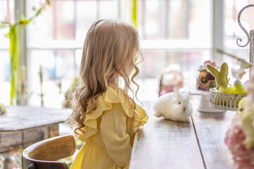A little blonde girl in a yellow dress is sitting at a festive Easter table with rabbits.A baby and a rabbit. The concept of Easter.Easter at the table. The Christian tradition.