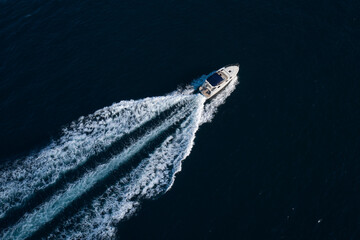 A white boat with a blue awning moving fast diagonally on dark blue water, top view.