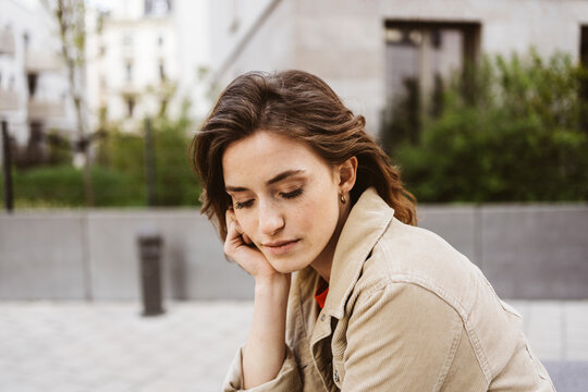 young woman sits pensively on a bench in the city and looks down