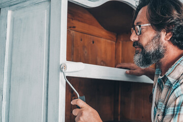 Close up of mature man painting and restoring an old antique wooden cabinet at home with roller and...