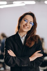 Attractive businesswoman in glasses posing with folded arms. Indoor shot of smiling young woman in black suit standing in office