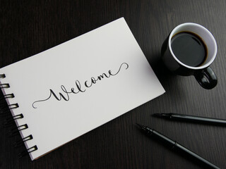 WELCOME lettering in notebook with cup of coffee and pens on black wooden desk