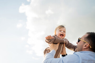 Dad playing with baby daughter laughing outdoors, happy people. The background of the sky is light.