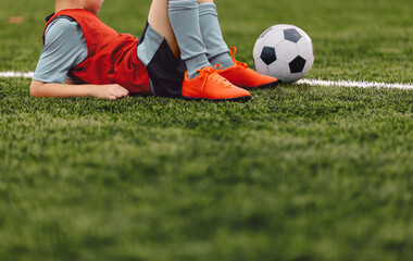 Young soccer boy with the ball lying and resting at soccer grass field at pitch sideline. Child at...