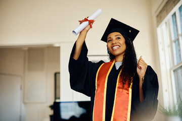 Happy black student holds her diploma while celebrating her graduation at home.
