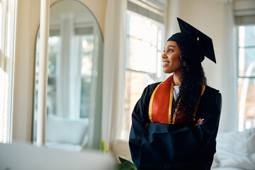 Confident African American female student in graduation gown at home.