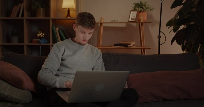 Handsome Young Man Using Laptop While Sitting on Sofa at Home. Male Millennial Person Browsing Internet and Looking at Computer Screen in Living Room. Concept of Working, Studying and Free Time.